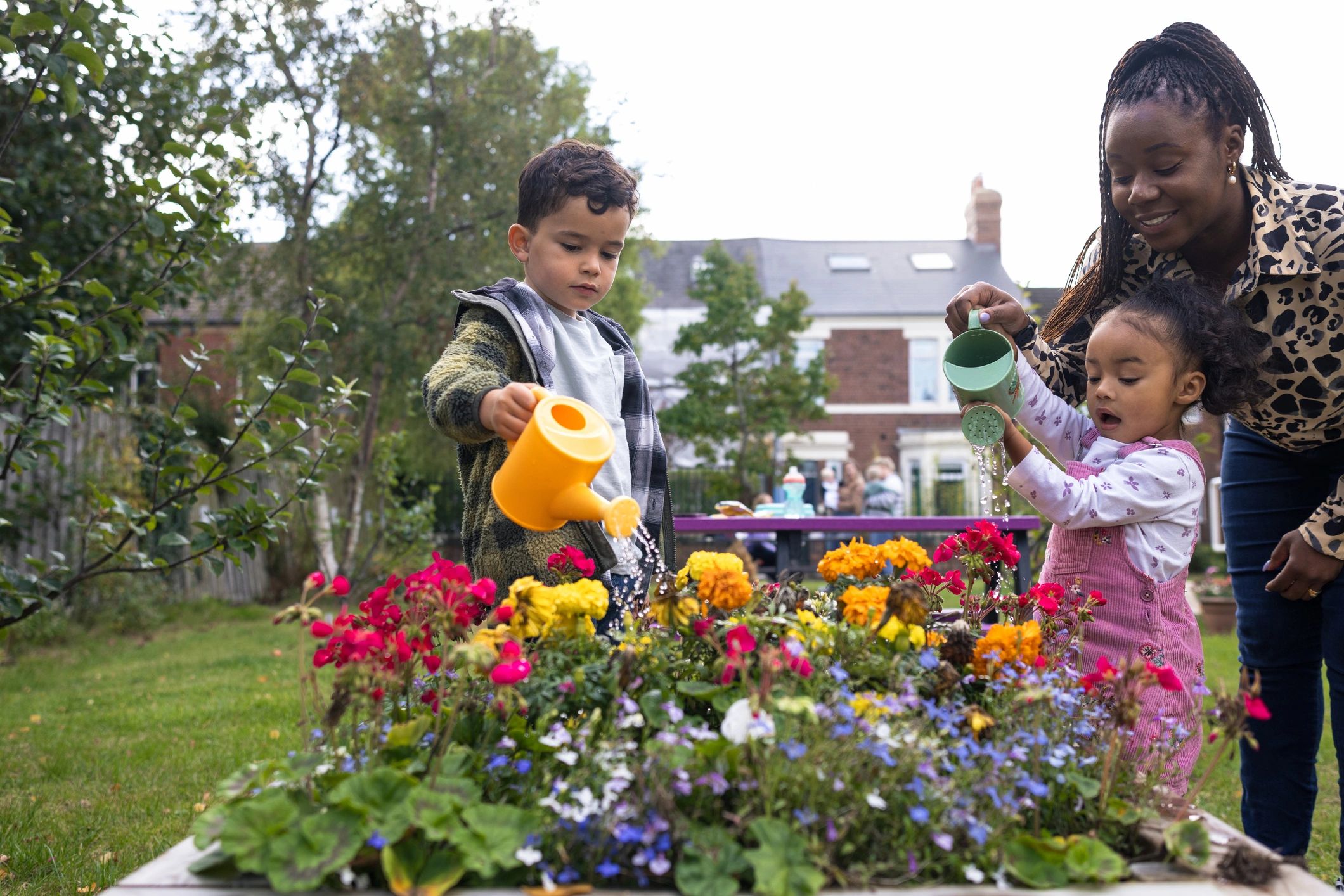 Children watering flowers in a garden while an adult looks on